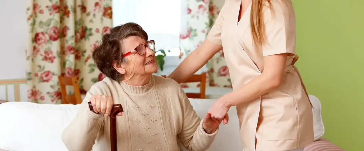 Betreuerin stützt eine ältere Dame beim Aufstehen im Wohnzimmer